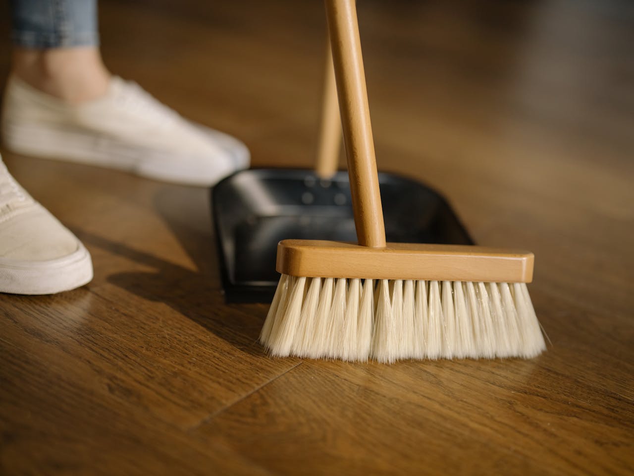 services-03 Close-up of a broom and dustpan with white sneakers indoors, representing housekeeping and cleanliness.